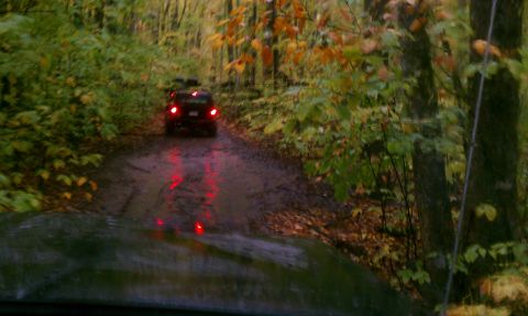 1979 Ford Bronco - SUV drives down a wet, muddy road surrounded by trees with fall foliage.