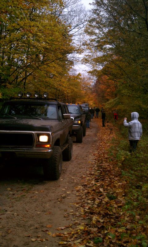 1979 Ford Bronco - A convoy of off-road vehicles, including a 1979 Ford Bronco, travels down a dirt path lined with autumn trees.