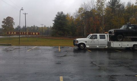 1979 Ford Bronco - A white flatbed truck transports a black pickup truck in front of the Dam Site Inn on a cloudy day.