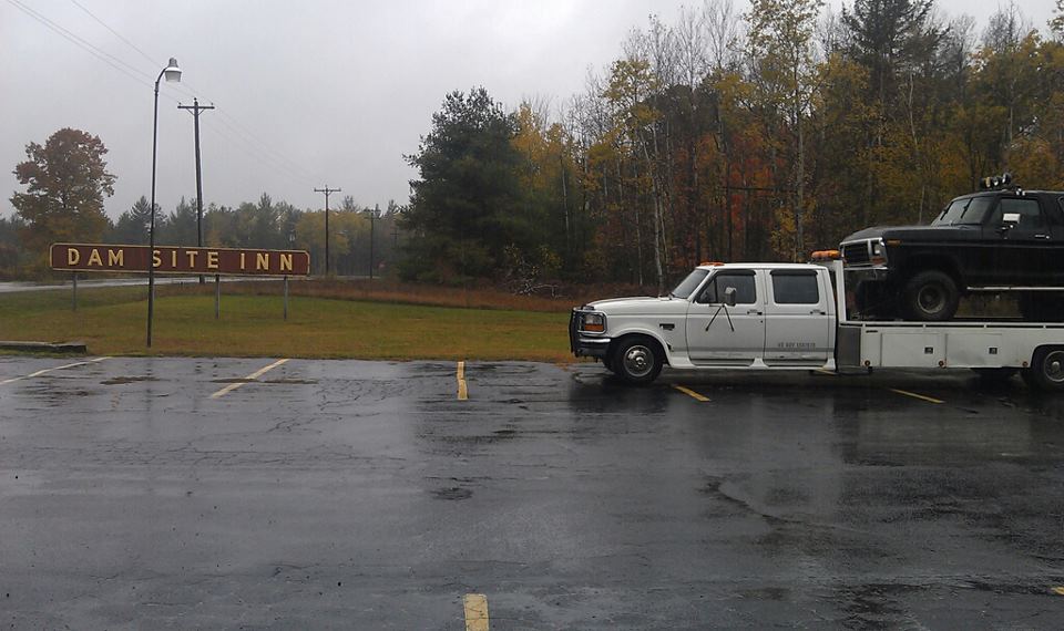 White flatbed truck carrying a black pickup truck, parked on a wet lot near a sign for Dam Site Inn