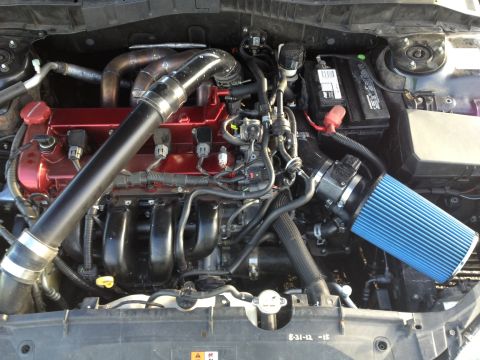 1987 Ford Bronco - A close-up view of a modified car engine bay, featuring a red valve cover, a large black intake pipe, and a blue cone air filter.