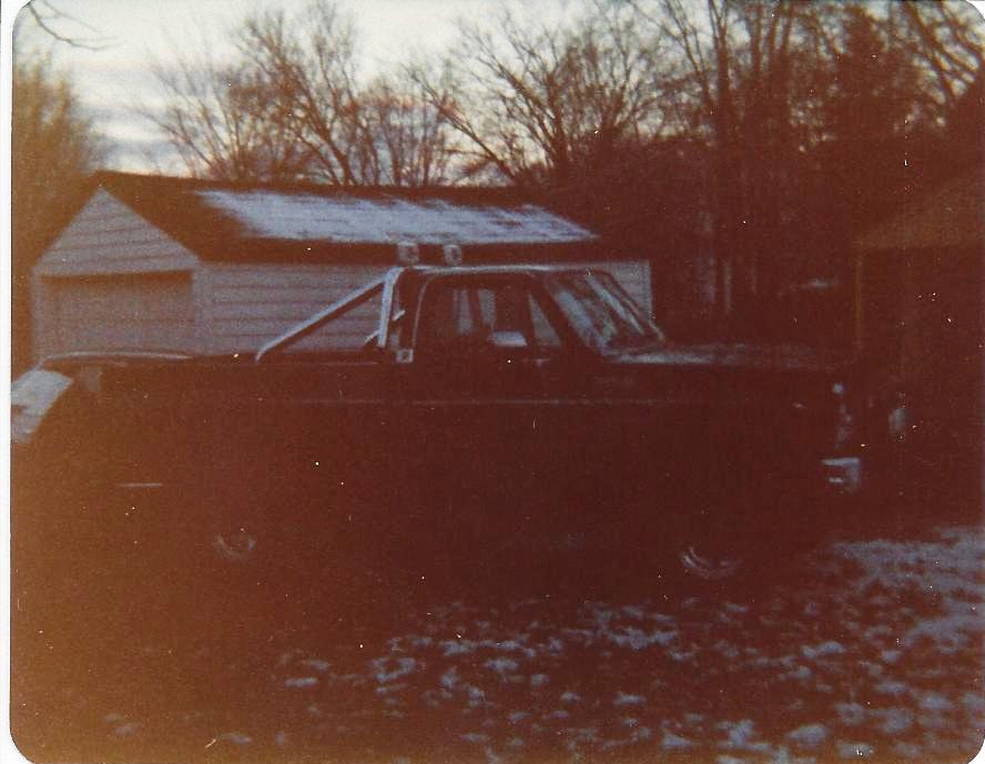 Dark pickup truck with roll bar and two lights, parked in a snowy yard next to a shed