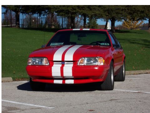 1990 Ford Mustang - A red 1990 Ford Mustang with iconic white racing stripes is parked in a lot, viewed from the front.