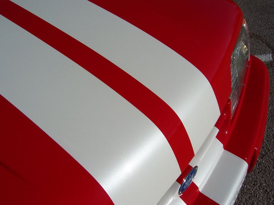 Red car hood with white racing stripes, showing part of the headlight and a Ford emblem