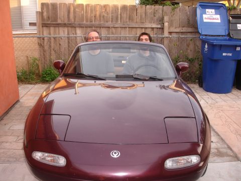 1993 Ford Taurus - A maroon Mazda Miata with pop-up headlights is parked in a driveway with two people visible inside.