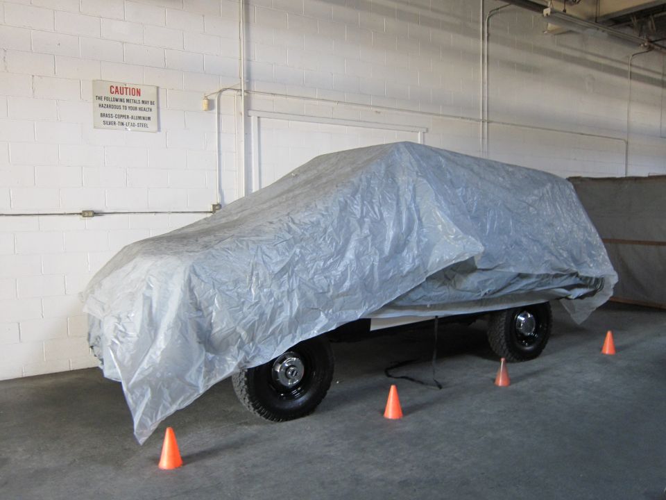 Vehicle covered with a gray tarp, parked indoors with orange cones on the floor