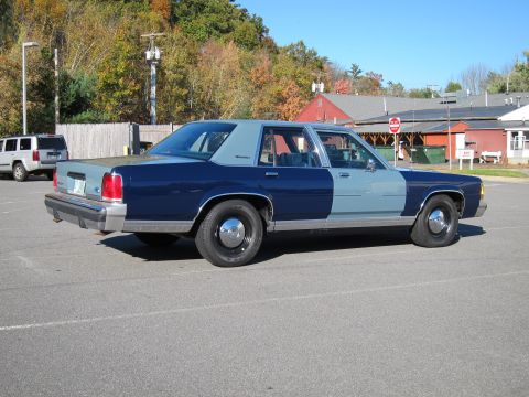 1991 Ford LTD Crown Victoria - A two-tone sedan with a flat front tire is parked in a lot with other vehicles and buildings in the background.