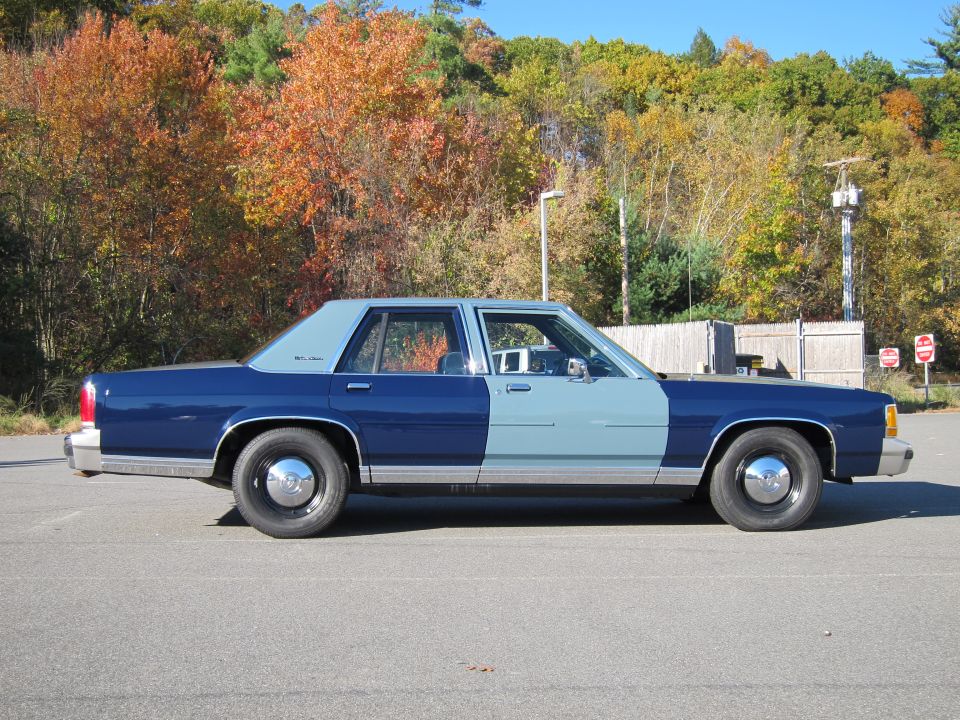 Side view of a blue and light blue sedan parked on asphalt with autumn trees in the background