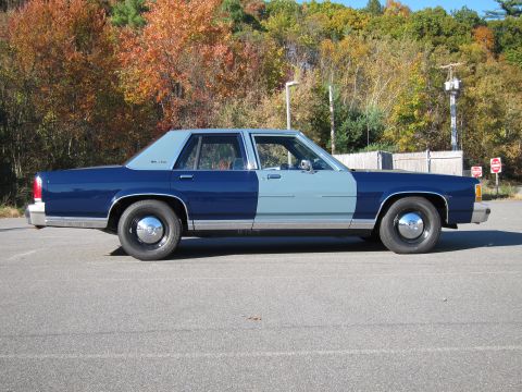 1991 Ford LTD Crown Victoria - A two-tone blue sedan with chrome hubcaps is parked on an asphalt lot with autumn trees in the background.