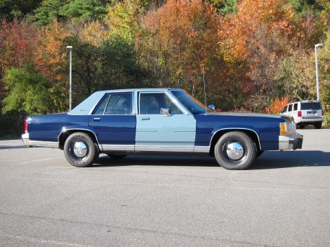 1991 Ford LTD Crown Victoria - A two-tone Ford Crown Victoria is parked in a lot with fall foliage behind it, and another SUV is visible in the distance.