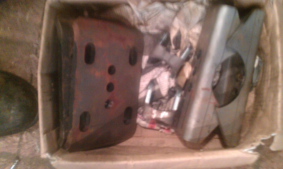 Two car parts, one rusty and rectangular with holes, the other silver and angular, sit in a cardboard box