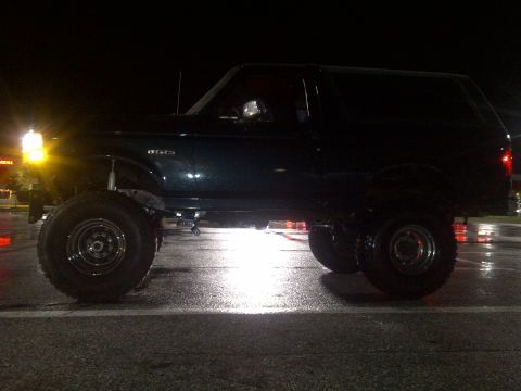 1995 Ford Bronco - Side view of a lifted 1995 Ford Bronco at night, showing its large tires and suspension modifications.