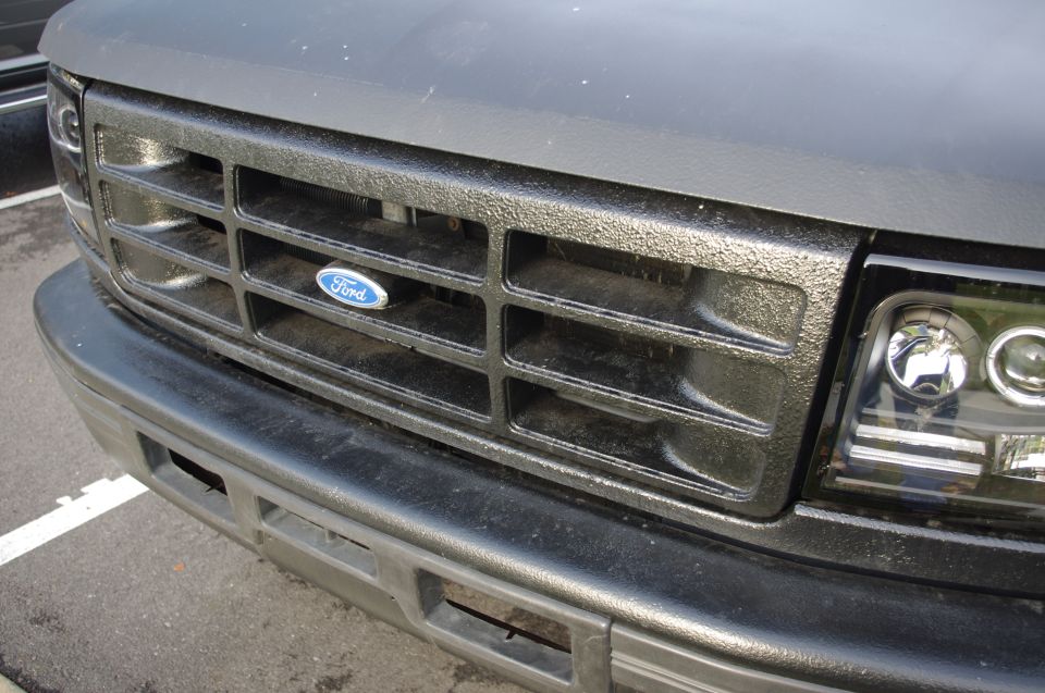 Close-up of a black Ford truck grille with a blue Ford oval badge. Headlight visible on the right