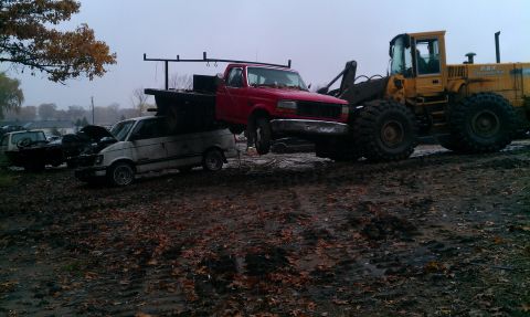 1995 Ford Bronco - A red pickup truck is being lifted by a yellow loader, with a white van and other vehicles in the background.