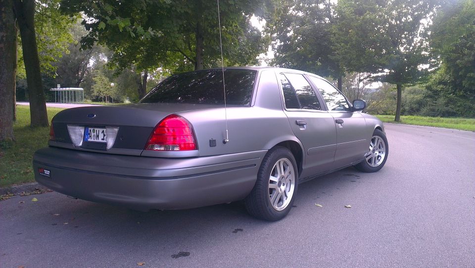 Gray Ford Crown Victoria parked on a road with trees in the background. License plate is visible