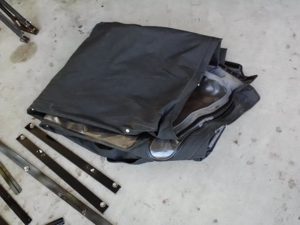 Folded black soft top with clear plastic windows and metal mounting strips on concrete floor