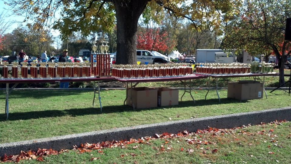 Rows of trophies on tables in a park setting with trees and parked vehicles in the background