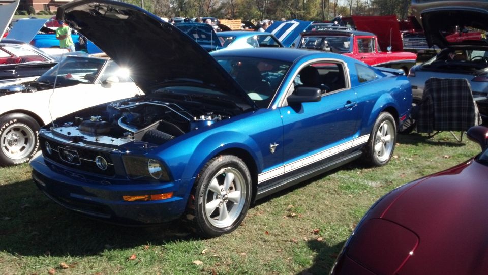 Blue Ford Mustang with hood open, parked on grass at a car show. Other cars visible