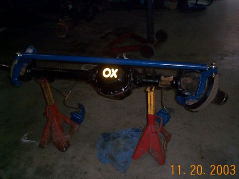 1977 Ford Bronco - A rebuilt front solid axle assembly rests on red jack stands in a garage, with the housing painted black, knuckles and tie rod painted blue…