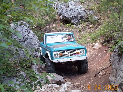 1977 Ford Bronco - Weathered turquoise early Ford Bronco with a white grille, roll bar and no doors driving down a narrow dirt trail between limestone outcrop…