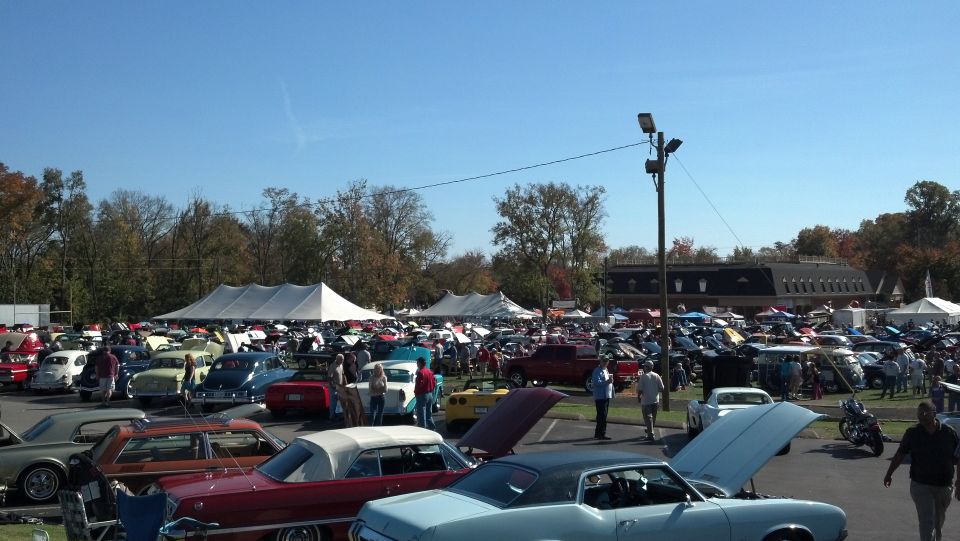 Outdoor car show with many vehicles parked under tents and in open areas. People are walking around looking at the cars