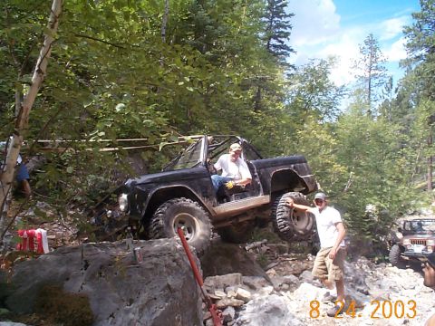 1977 Ford Bronco - A flat-black topless early Ford Bronco with an interior roll bar and cut rear fenders crawls a rocky wooded trail with one rear wheel lifte…