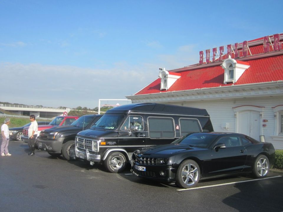 Several vehicles parked in a lot, including a black van and a black coupe. A building with a red roof is in the background