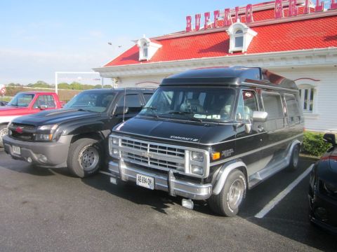 1991 Ford LTD Crown Victoria - A black Starcraft conversion van is parked in a lot, with other vehicles nearby and a building with a red roof in the background.