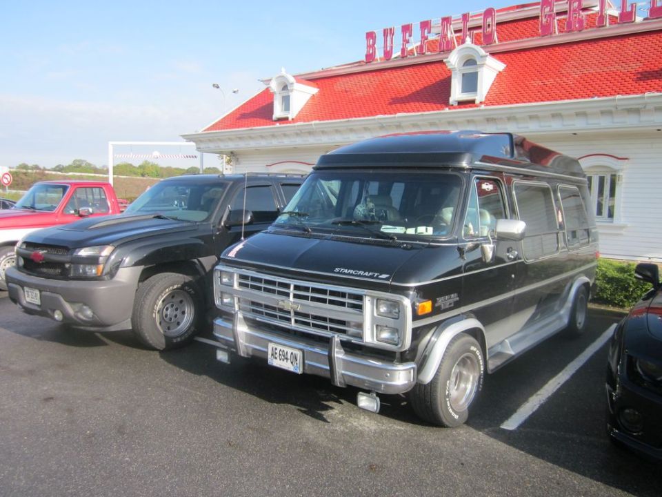 Black conversion van parked next to a dark SUV and a red pickup truck in front of a building with a red roof