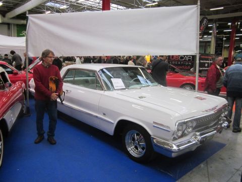 1991 Ford LTD Crown Victoria - A white 1963 Chevrolet Impala is displayed indoors with a man standing beside it and other vehicles in the background.