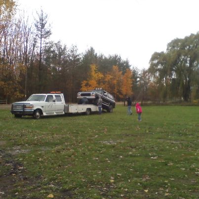 1979 Ford Bronco - Tow truck transporting a dark SUV in a field with trees in the background.