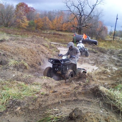 1979 Ford Bronco - ATVs tackle a challenging muddy terrain, with a dark SUV following in the background under an overcast sky.