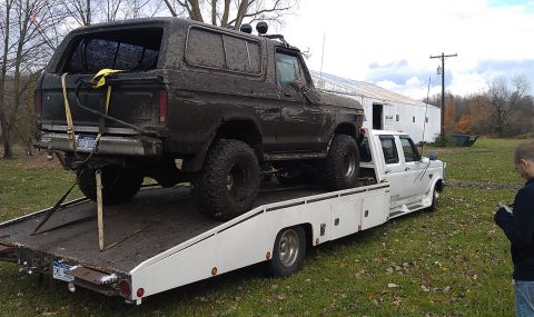 1979 Ford Bronco - A heavily muddied 1979 Ford Bronco is loaded onto a white flatbed tow truck, suggesting it was recently recovered from off-road conditions.