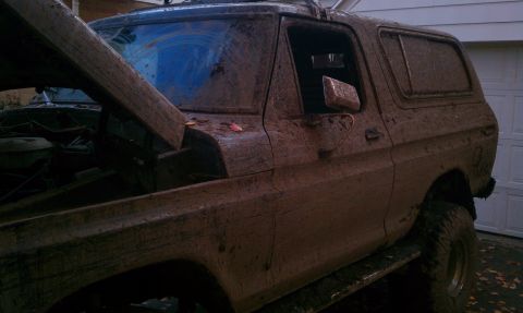 1979 Ford Bronco - This heavily mud-covered SUV, likely a 1978 Ford Bronco, is parked with its hood up in front of a garage.