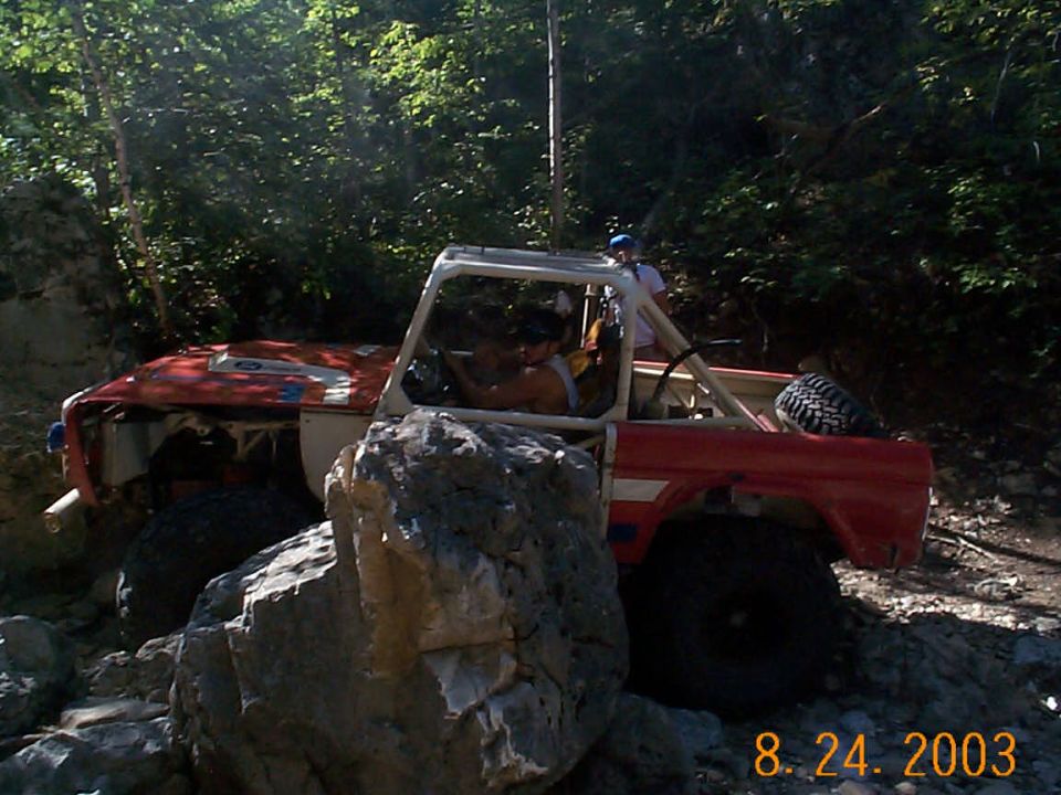 Red off-road vehicle with a roll cage navigating a rocky trail with trees in the background
