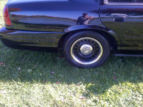 2001 Ford Crown Victoria - A black sedan sits on a grassy area, showing its rear wheel and part of the body.