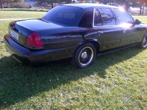 2001 Ford Crown Victoria - A dark-colored sedan is parked on a grassy area, with the sun glinting off its windows.