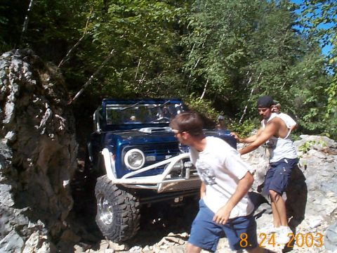 1977 Ford Bronco - A blue off-road SUV is being guided through a narrow, rocky path by two men.