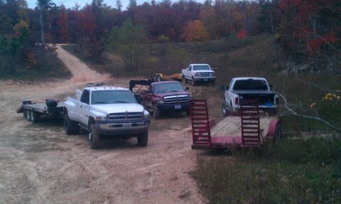 1987 Ford Bronco - Multiple trucks and trailers are parked on a dirt path surrounded by trees with fall colors.