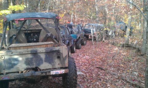 1987 Ford Bronco - Several off-road vehicles are parked on a leaf-covered trail in a wooded area during autumn.