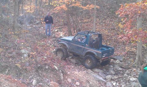 1987 Ford Bronco - A person drives a modified off-road vehicle through a wooded area with fallen leaves on the ground.