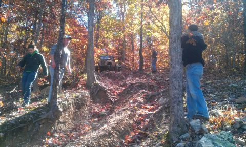 1987 Ford Bronco - People watch an off-road vehicle navigate a challenging trail in a forest during fall.