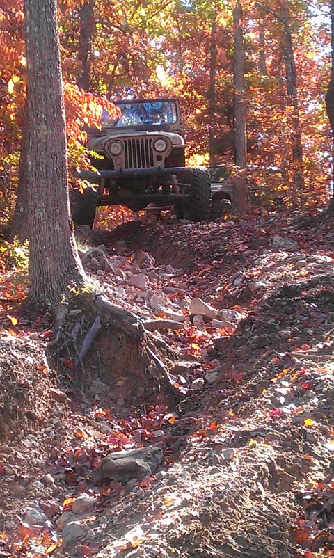 1987 Ford Bronco - A tan Jeep CJ-5 navigates a challenging, rocky trail surrounded by trees in autumn foliage.