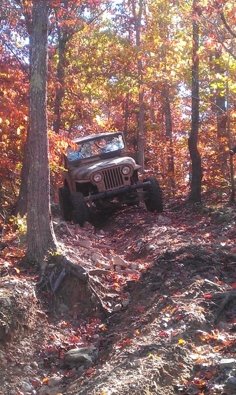 1987 Ford Bronco - A brown Jeep CJ-5 navigates a challenging rocky trail surrounded by trees with colorful autumn foliage.