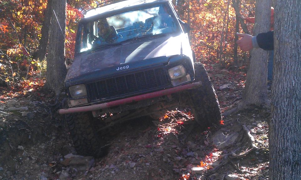 Jeep Cherokee XJ climbing a rocky, wooded trail with people watching