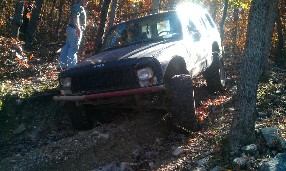 Jeep Cherokee off-roading on a dirt trail, front 3/4 view, dark color, mud-splattered