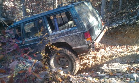 1987 Ford Bronco - A dark green Jeep Cherokee XJ tackles a challenging off-road trail with steep inclines and rocky obstacles.