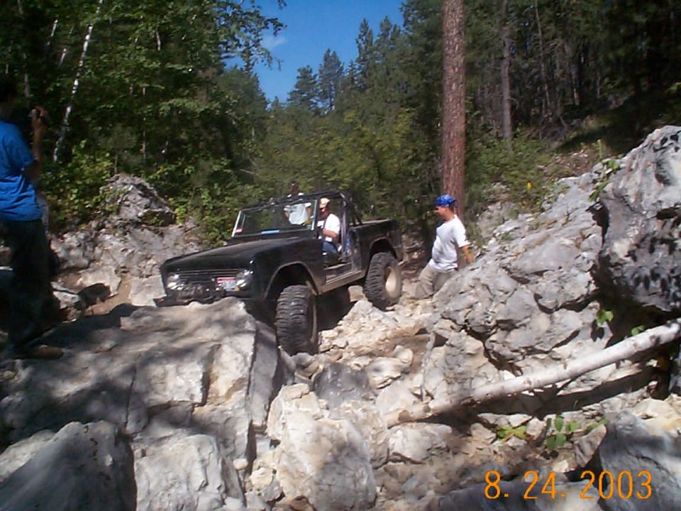 1977 Ford Bronco navigating a steep, rocky trail with two people inside and one person standing nearby