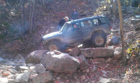 1987 Ford Bronco - A Jeep Cherokee XJ tackles a rocky off-road course with people watching.