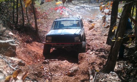1987 Ford Bronco - A Jeep Cherokee XJ navigates a challenging off-road trail with muddy terrain and trees.